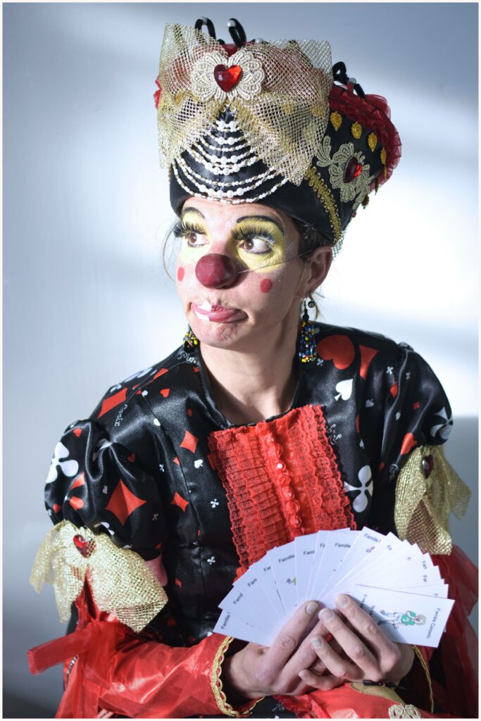 Un clown vêtu d’un costume noir et rouge à motifs de cartes à jouer tient un éventail de cartes blanches dans ses mains. Son maquillage expressif, avec un nez rouge, des joues ornées de ronds rouges et un fard doré autour des yeux, accentue son regard malicieux. Elle porte une coiffe majestueuse ornée de perles, de dentelle dorée et de cœurs rouges, renforçant son allure royale et fantaisiste. Son expression facétieuse et son regard en coin laissent deviner une surprise ou une plaisanterie à venir. L’éclairage crée un contraste entre lumière et ombre, mettant en valeur les détails de son costume et de son visage.