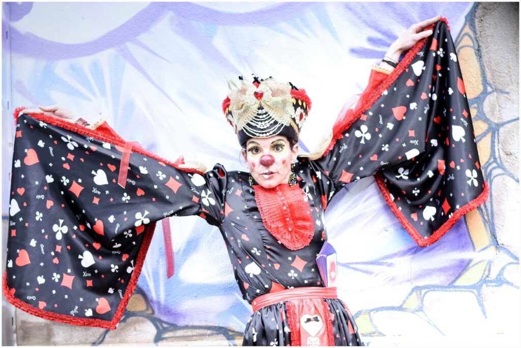 Un clown extravagant vêtu d'un costume inspiré de la Dame de Cœur, noir et rouge avec des motifs de cartes à jouer, pose avec les bras grands ouverts et une expression théâtrale de surprise. Son maquillage coloré, avec un nez rouge et des joues décorées de cœurs, renforce son allure fantaisiste. Des cartes flottent autour d'elle, donnant une impression de magie et de mouvement. En arrière-plan, une fresque murale représente un visage aux cheveux bleus, ajoutant une touche artistique et dynamique à la scène.