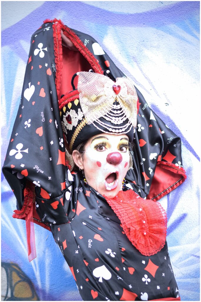 Un clown extravagant vêtu d'un costume inspiré de la Dame de Cœur, noir et rouge avec des motifs de cartes à jouer, pose avec les bras grands ouverts et une expression théâtrale de surprise. Son maquillage coloré, avec un nez rouge et des joues décorées de cœurs, renforce son allure fantaisiste.